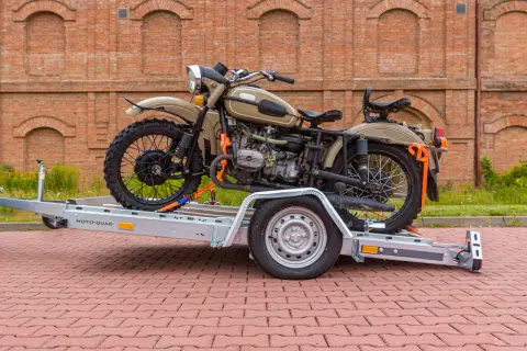Motocykl Ural na przyczepie z pasami transportowymi, na tle ceglanego budynku.